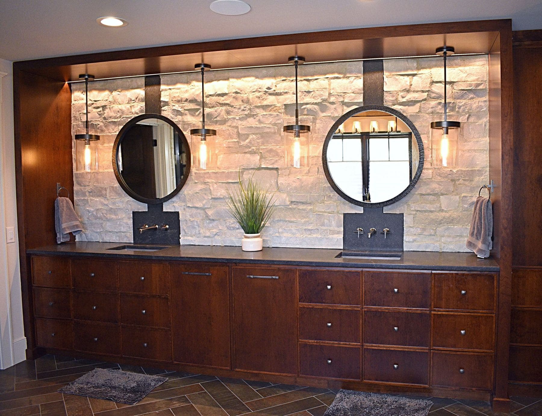 Dark stained wood double vanity with many drawers against a limestone wall, with two round mirrors and pendant lights
