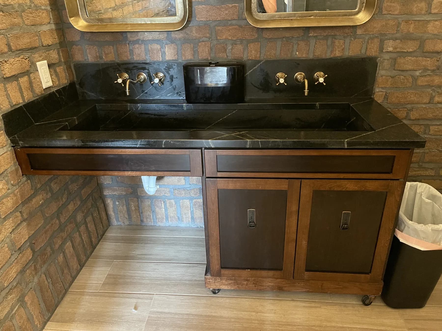 Black stone vanity top with wall-mounted brass faucets on brick