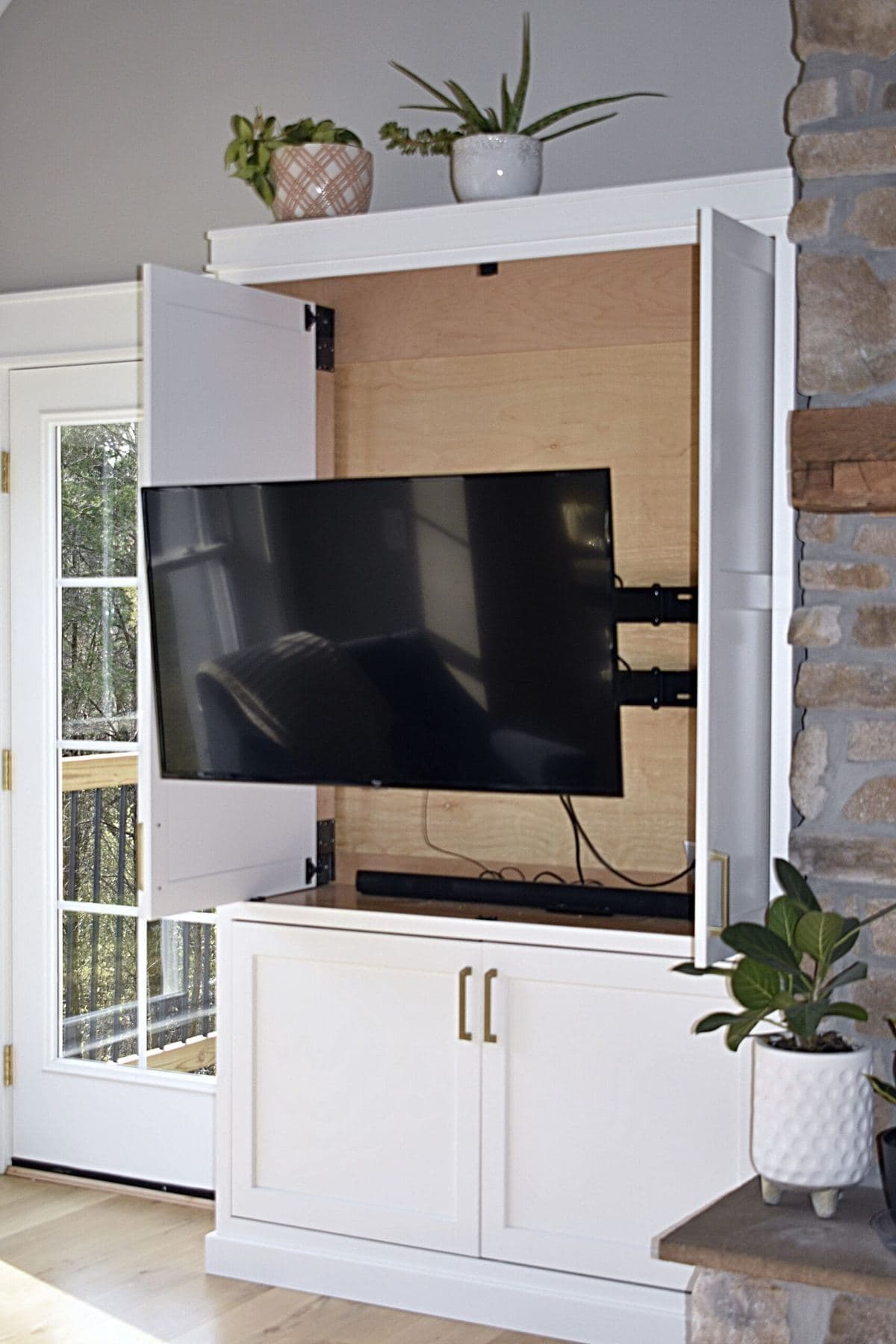 White built-in cabinet with bi-fold doors open to reveal a wall-mounted television, beside a stone fireplace