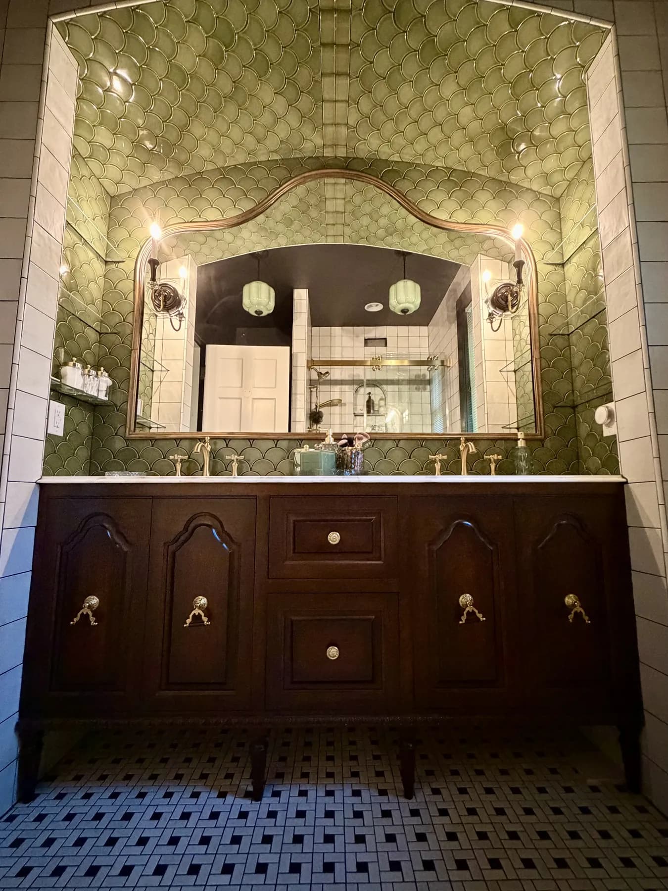 Stained double vanity under an arched mirror in a green tiled bath