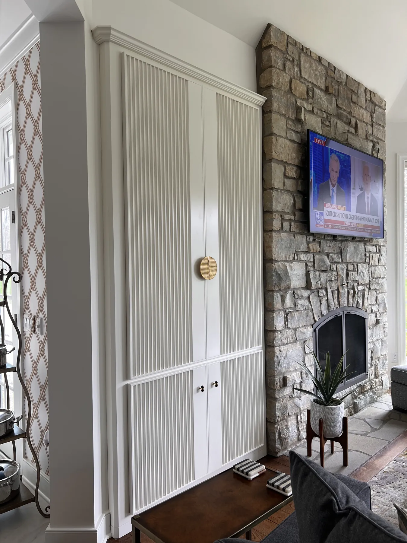 Reeded painted cabinet beside a stone fireplace with a wall-mounted television
