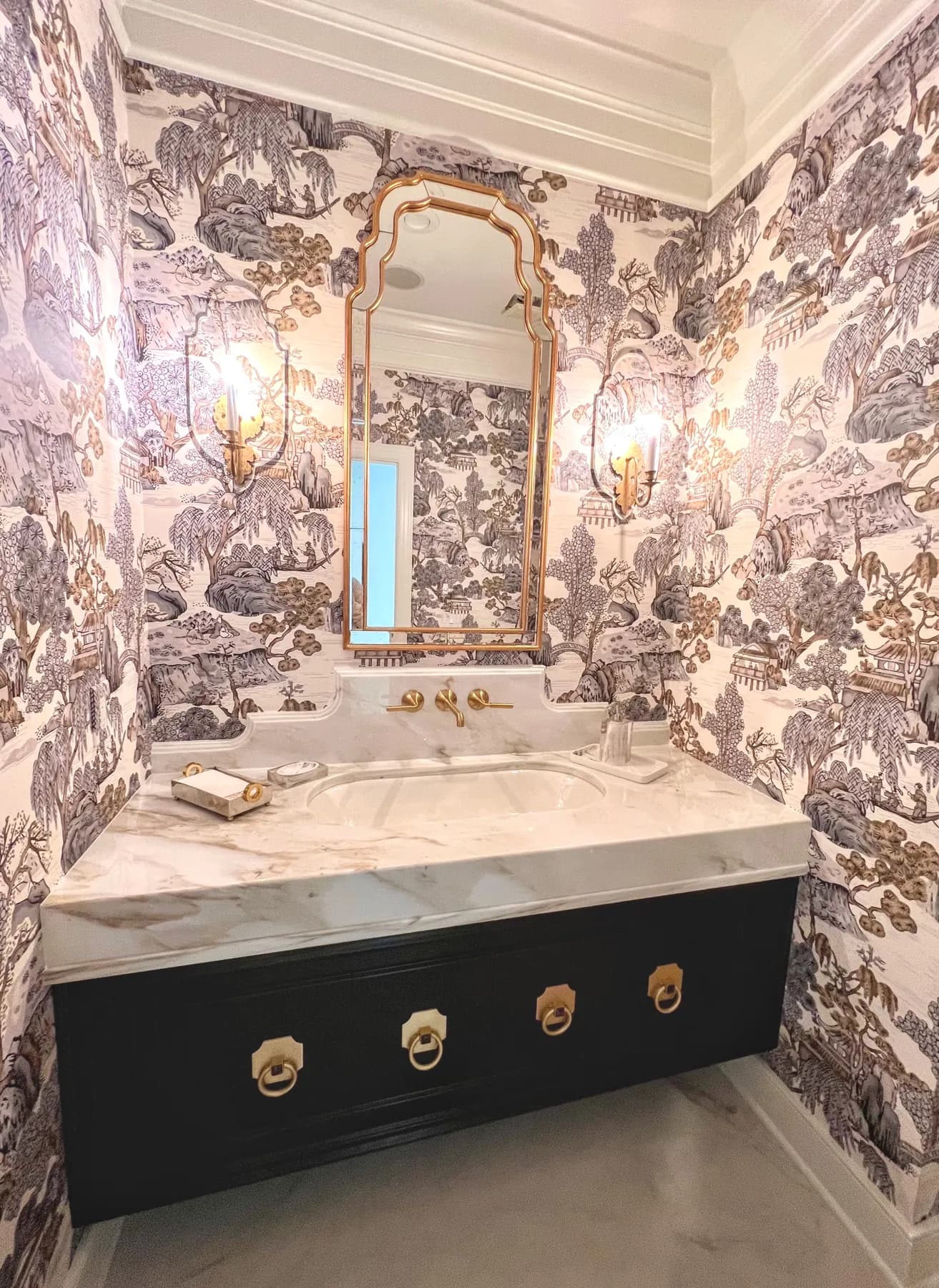 Black vanity with brass ring pulls under a marble top in a toile papered powder room