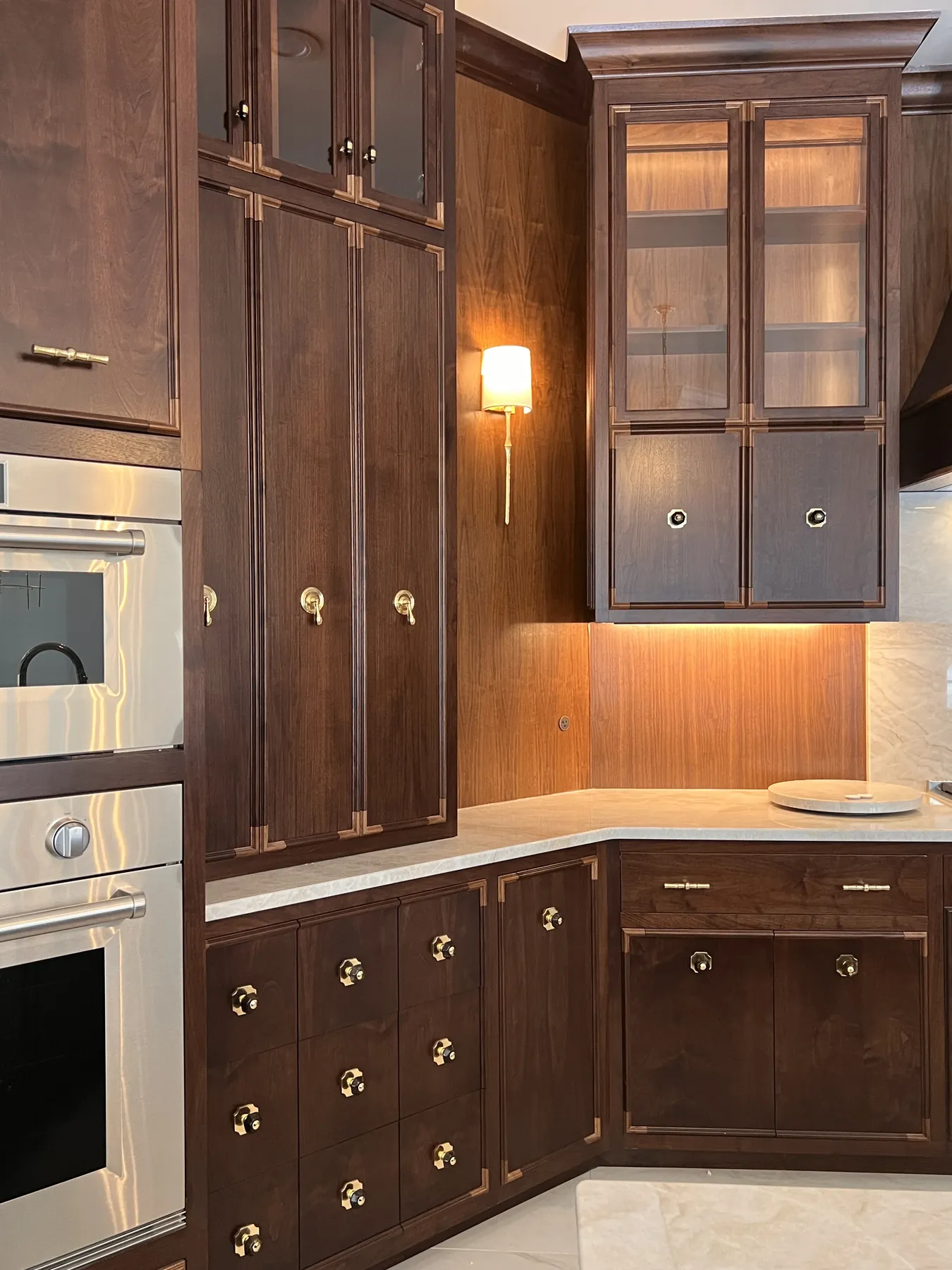 Walnut cabinetry around integrated wall ovens with a lit sconce and glass uppers