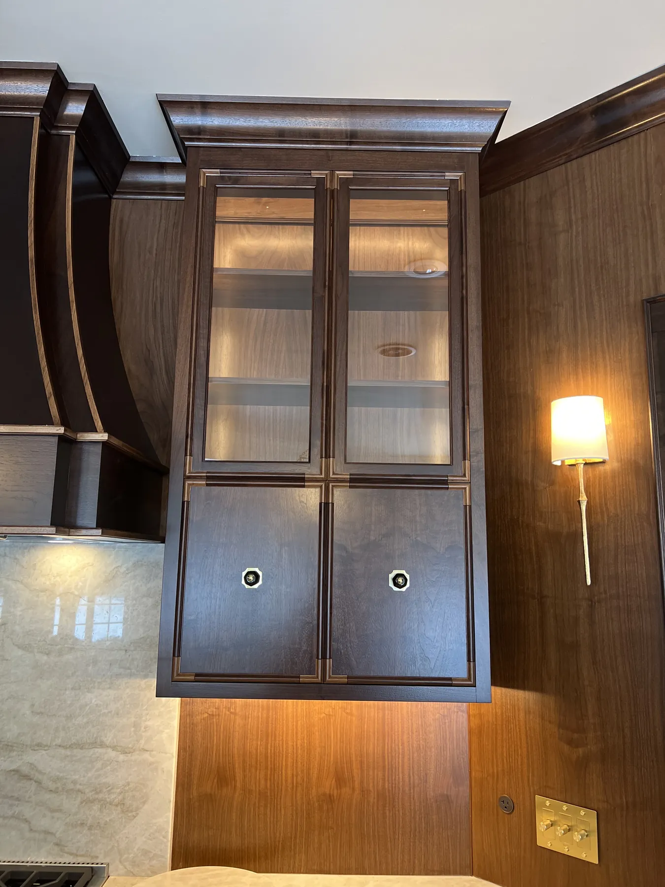 Glass-front walnut wall cabinet lit from within beside a brass sconce