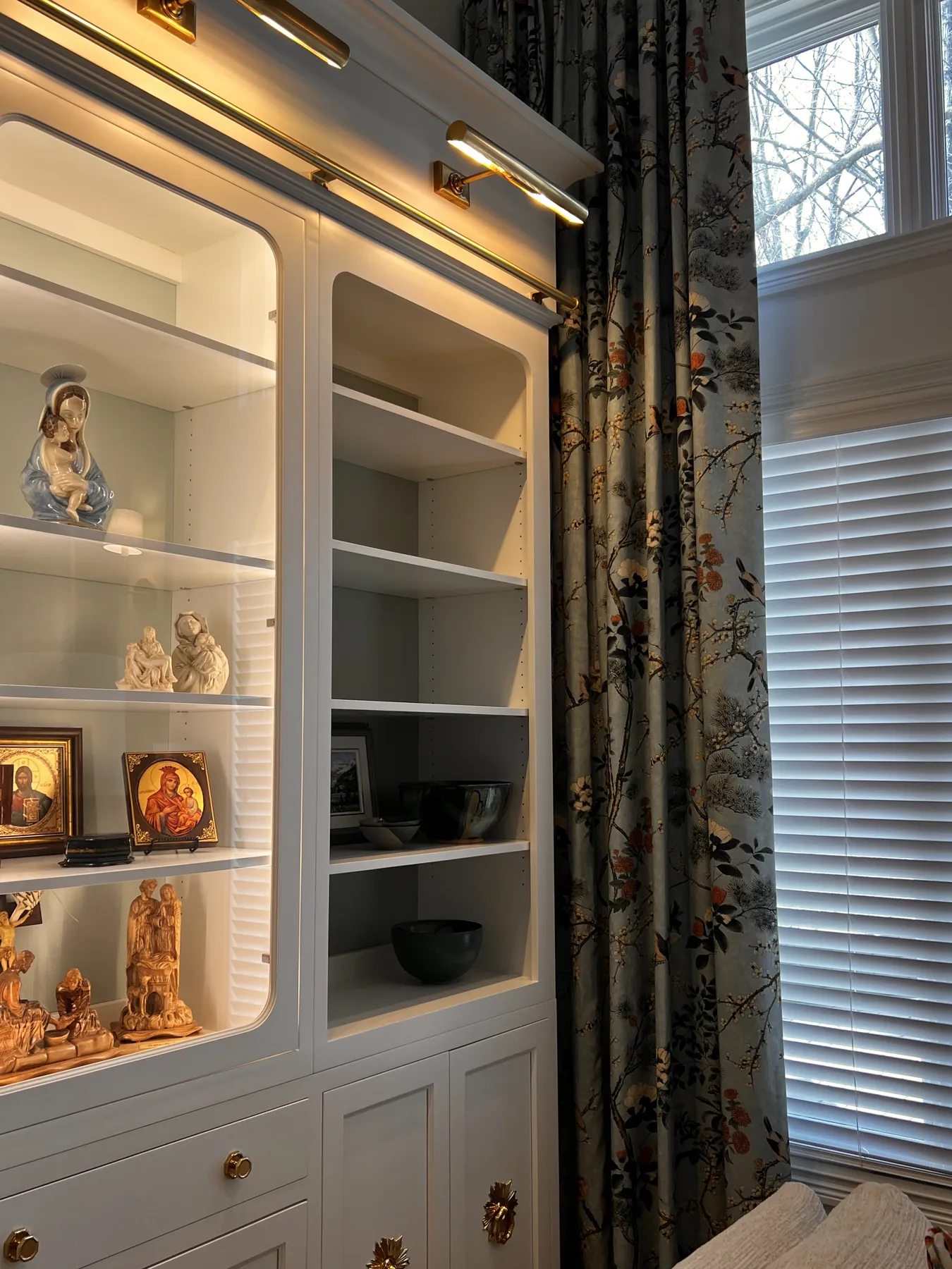 Lit painted shelves holding pottery beside a curtained window